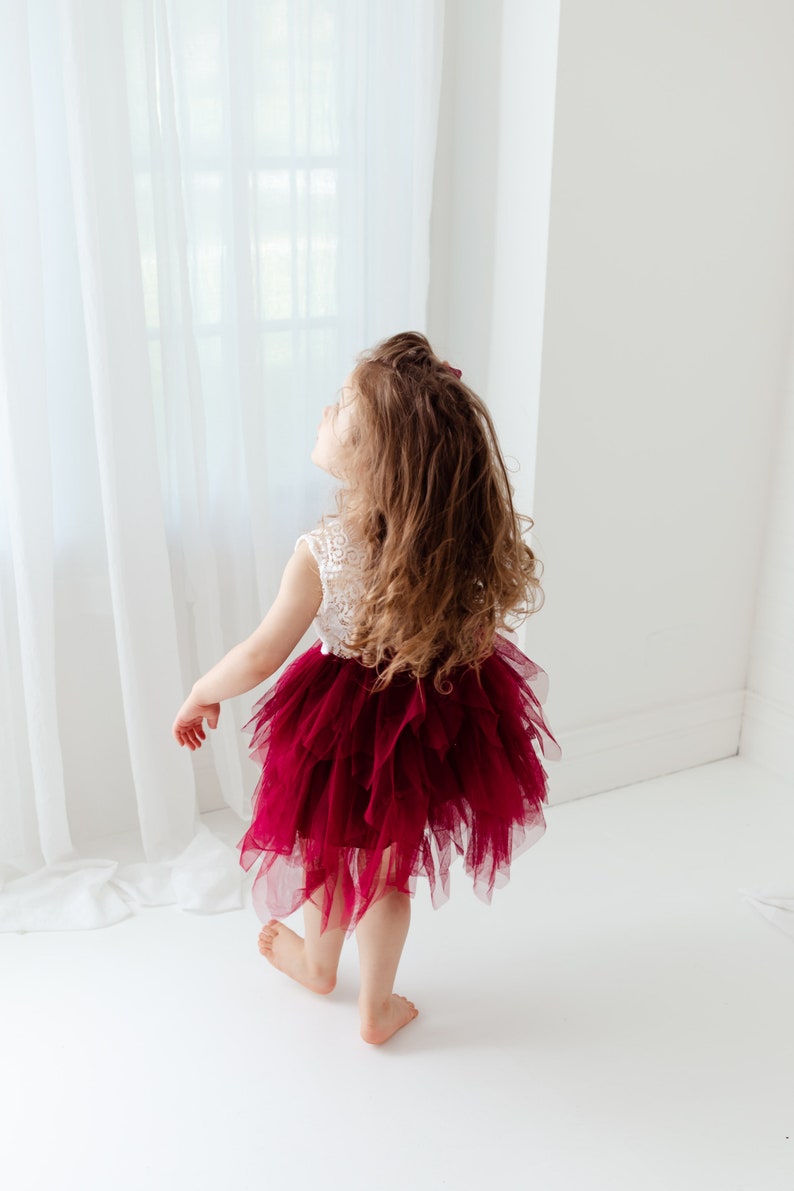 Puede incluir: Una ni&ntilde;a con un top de encaje blanco y una falda de tul rojo oscuro en capas. La ni&ntilde;a mira hacia arriba, con cabello casta&ntilde;o largo y suelto. El vestido contrasta vivamente con el fondo blanco.