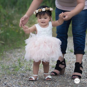 May include: A young girl in a pink tulle dress and a flower crown is being led by an adult wearing jeans and a light pink shirt. The girl is smiling and looking at the camera.
