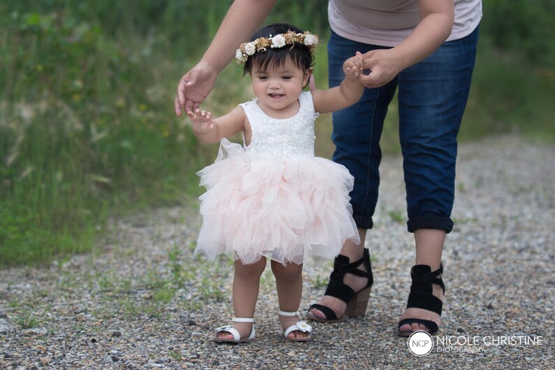 May include: A young girl in a pink tulle dress and a flower crown is being led by an adult wearing jeans and a light pink shirt. The girl is smiling and looking at the camera.