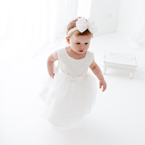 Puede incluir: Una ni&ntilde;a peque&ntilde;a con un vestido blanco con un corpi&ntilde;o de encaje y una diadema blanca con un lazo. El vestido tiene una falda de tul y est&aacute; en capas. La ni&ntilde;a est&aacute; de pie sobre una superficie blanca.
