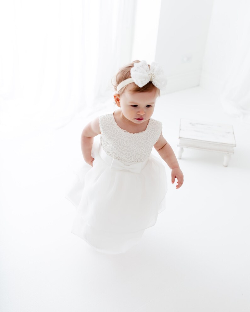Puede incluir: Una ni&ntilde;a peque&ntilde;a con un vestido blanco con un corpi&ntilde;o de encaje y una diadema blanca con un lazo. El vestido tiene una falda de tul y est&aacute; en capas. La ni&ntilde;a est&aacute; de pie sobre una superficie blanca.