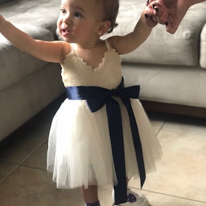 May include: A little girl wearing a white tulle dress with a navy blue sash and white shoes. She is holding hands with someone off camera.