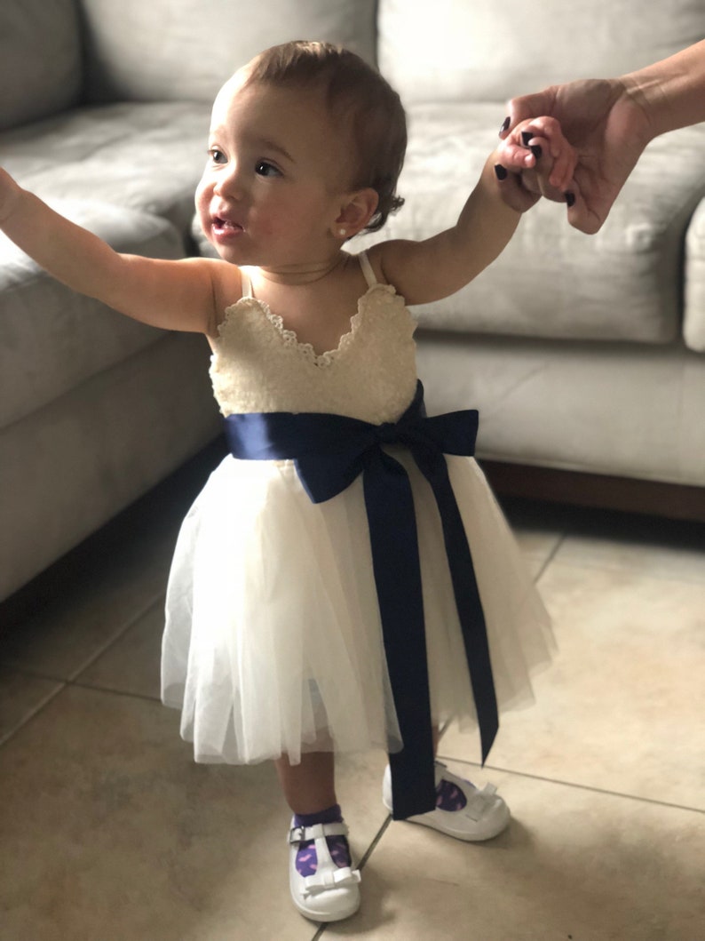 May include: A little girl wearing a white tulle dress with a navy blue sash and white shoes. She is holding hands with someone off camera.