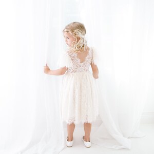 May include: A cream-colored dress featuring a lace bodice with short flutter sleeves and a scalloped hemline. The dress has a V-shaped back and a layered skirt. A gold-toned floral headpiece with white ribbons is worn on the head, and simple white flat shoes are worn on the feet.