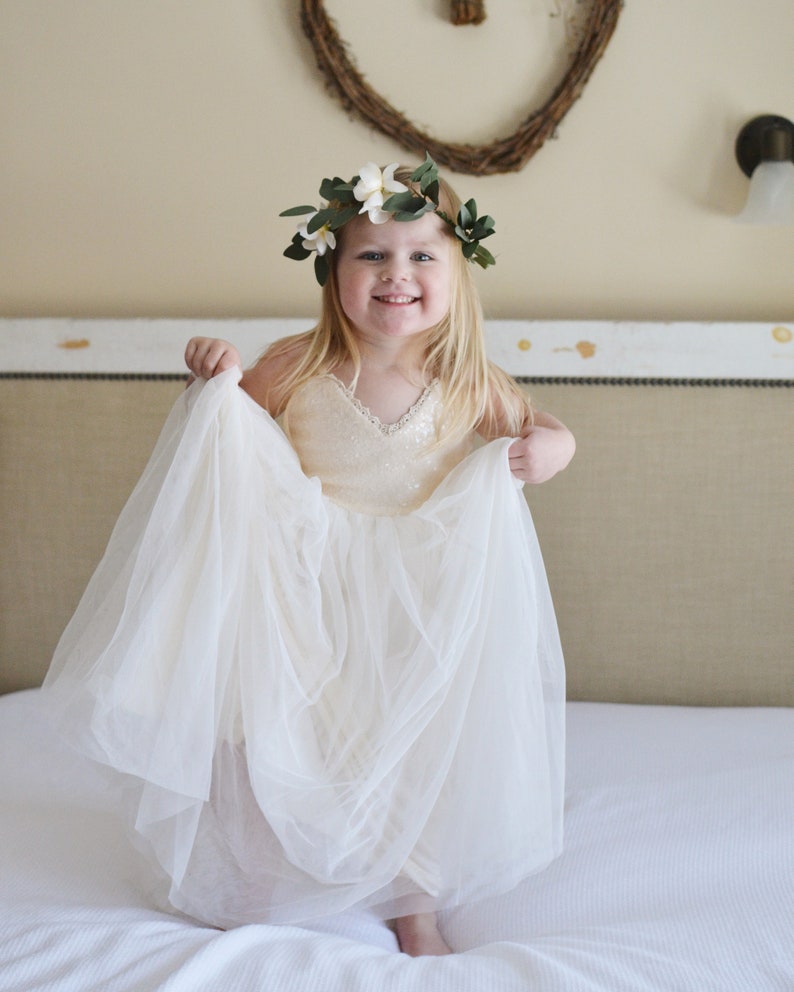 May include: A young girl wearing a white tulle dress and a flower crown with white flowers and green leaves. She is smiling and holding the skirt of her dress.
