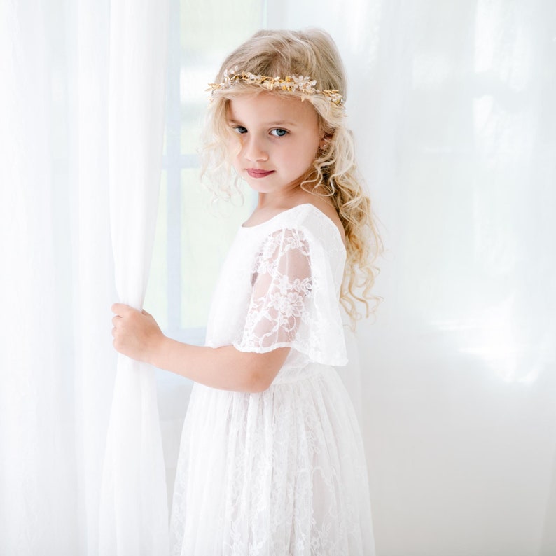 May include: A young girl wearing a white lace dress with a gold leaf crown. She is standing next to a white curtain and looking at the camera.