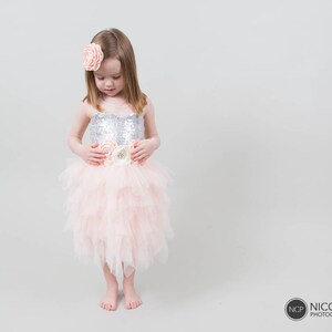 May include: A young girl wearing a pink tulle dress with a silver sequined bodice and a floral embellishment at the waist. She is wearing a pink flower hair clip.