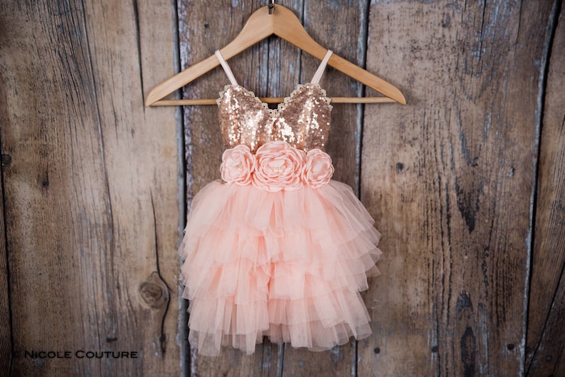 May include: A sleeveless dress featuring a rose gold sequined bodice with thin straps and a lace-trimmed neckline. The waist is adorned with three large peach-colored fabric flowers, and the skirt consists of multiple layers of ruffled peach tulle.
