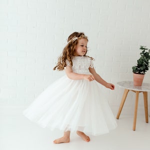 May include: A young girl wearing a white tulle dress with a lace bodice and a gold flower crown. She is standing on a white surface and is twirling her dress.