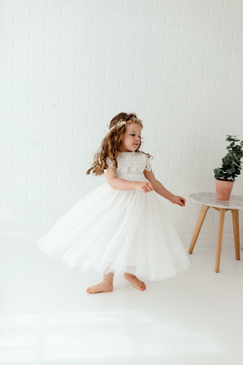 May include: A young girl wearing a white tulle dress with a lace bodice and a gold flower crown. She is standing on a white surface and is twirling her dress.