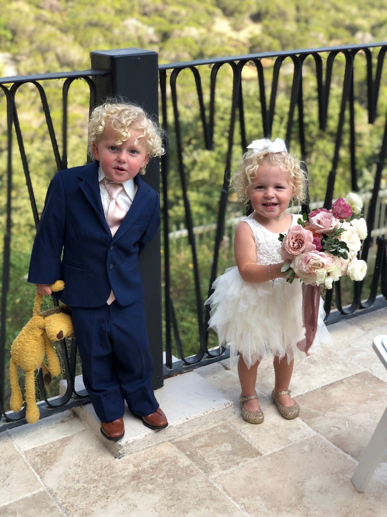 Op de afbeelding: Een jonge jongen in een donkerblauw pak houdt een knuffeldier vast terwijl een jong meisje in een witte jurk een boeket roze en witte bloemen vasthoudt.
