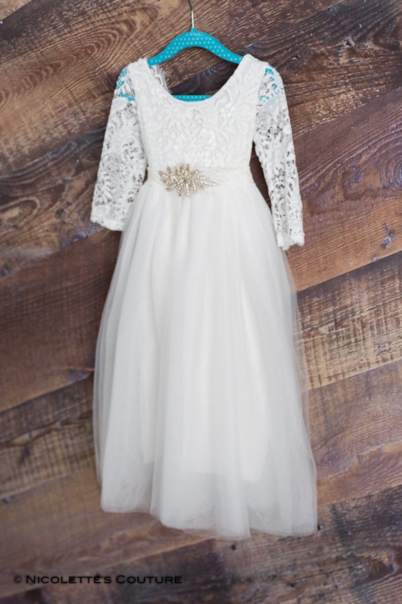 Puede incluir: Un vestido de tul blanco con mangas de encaje y un cintur&oacute;n de cuentas. El vestido est&aacute; colgado en una percha turquesa sobre un fondo de madera.