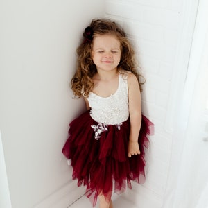 Puede incluir: Una ni&ntilde;a peque&ntilde;a con un vestido blanco y burdeos con una falda de tul en capas. El corpi&ntilde;o es blanco con adornos florales. Tiene el pelo casta&ntilde;o rizado y un accesorio para el pelo burdeos. La ni&ntilde;a est&aacute; de pie en una esquina con los ojos cerrados.