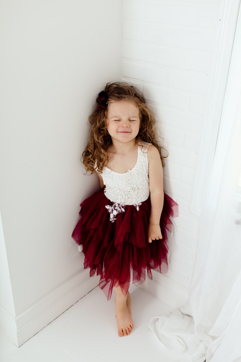 Puede incluir: Una ni&ntilde;a peque&ntilde;a con un vestido blanco y burdeos con una falda de tul en capas. El corpi&ntilde;o es blanco con adornos florales. Tiene el pelo casta&ntilde;o rizado y un accesorio para el pelo burdeos. La ni&ntilde;a est&aacute; de pie en una esquina con los ojos cerrados.