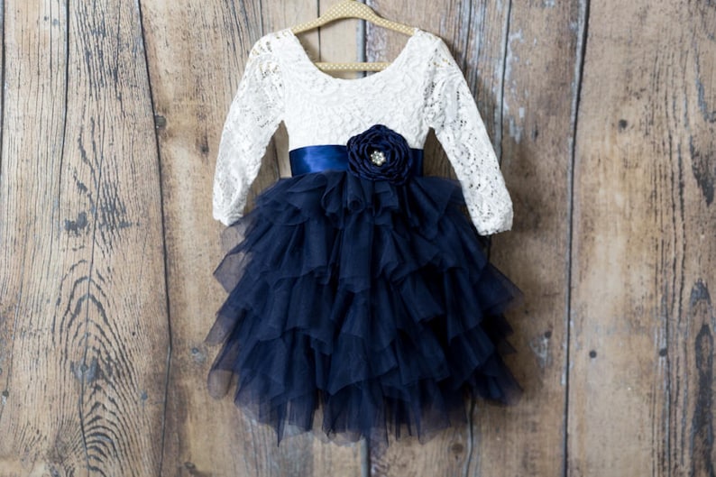 May include: A long-sleeved dress featuring a white lace bodice with a scoop neckline and a navy blue satin waistband. The skirt consists of multiple layers of navy blue tulle ruffles, accented by a large navy blue fabric flower with a pearl centre.