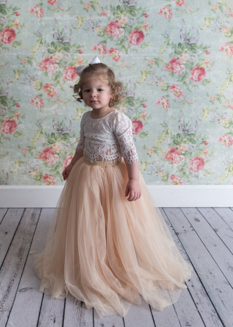 May include: A two-piece outfit consisting of a white lace long-sleeve top with a scalloped hem and a floor-length, voluminous peach-colored tulle skirt with an elastic waistband. A small white bow is attached to the hair.