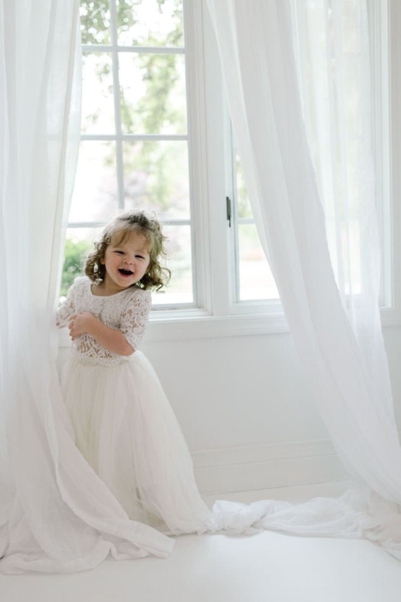 May include: A young child wearing a white lace top and a flowing tulle skirt, standing near a window with white curtains. The child is smiling and laughing, creating a joyful and innocent atmosphere. The dress has a delicate, elegant design.