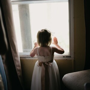 May include: A young girl in a white dress with a brown sash stands by a window, looking out. She has her hands raised and is wearing a white bow in her hair.