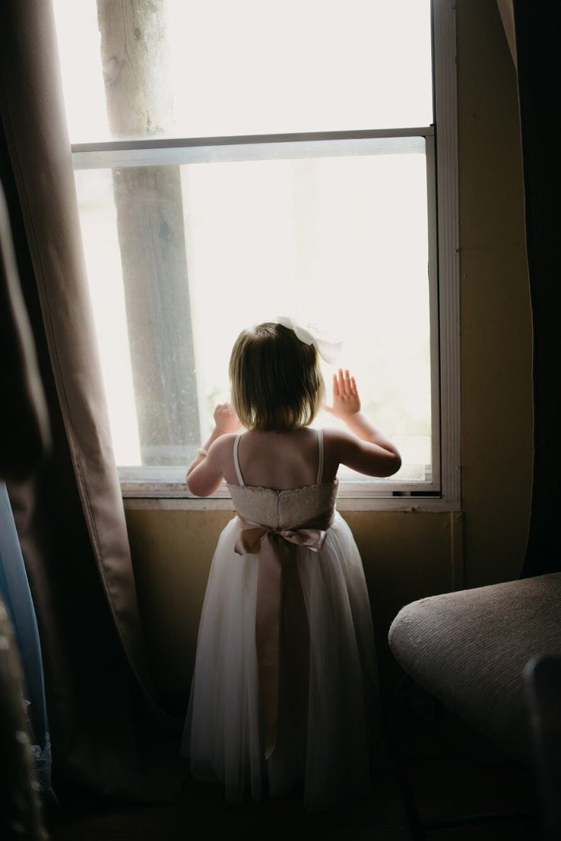 May include: A young girl in a white dress with a brown sash stands by a window, looking out. She has her hands raised and is wearing a white bow in her hair.