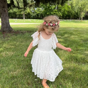 May include: A young girl wearing a white lace dress and a flower crown with pink and white flowers. She is standing in a grassy area with trees in the background.