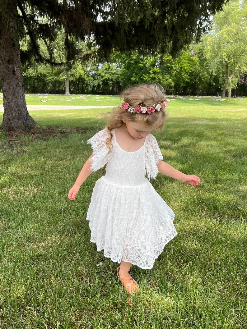 May include: A young girl wearing a white lace dress and a flower crown with pink and white flowers. She is standing in a grassy area with trees in the background.