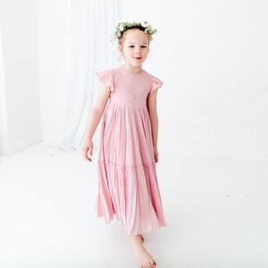 May include: A young girl wearing a pink, tiered, lace dress with a floral crown. She is standing in a white room and smiling at the camera.