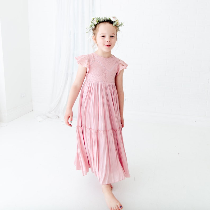 May include: A young girl wearing a pink, tiered, lace dress with a floral crown. She is standing in a white room and smiling at the camera.
