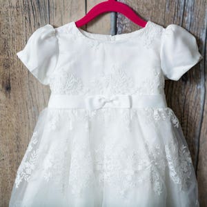May include: A white dress featuring short puff sleeves, a round neckline, and a satin waistband with a small centered bow. The bodice and skirt are adorned with intricate floral lace embroidery, and the skirt has a layered tulle overlay.