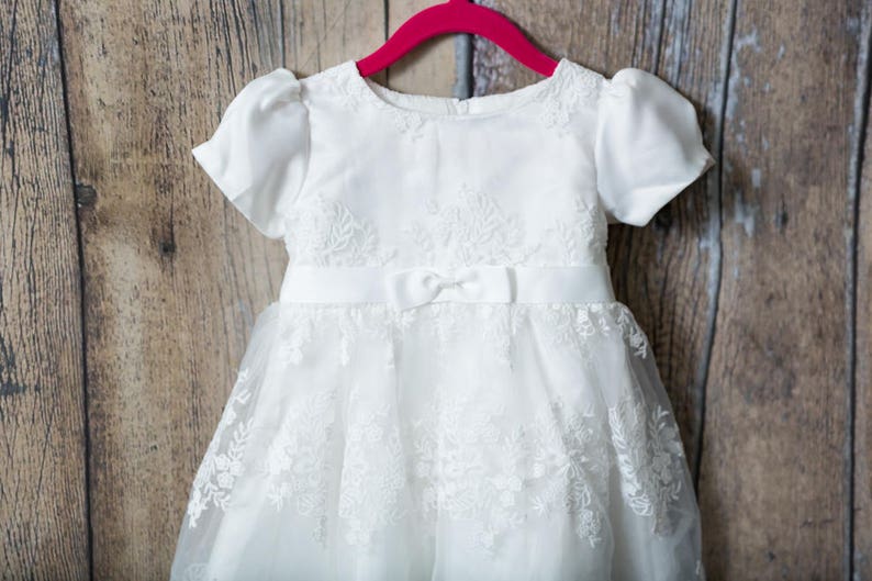 May include: A white dress featuring short puff sleeves, a round neckline, and a satin waistband with a small centered bow. The bodice and skirt are adorned with intricate floral lace embroidery, and the skirt has a layered tulle overlay.