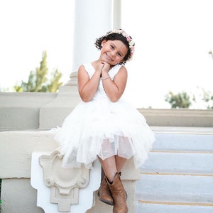 May include: A young girl wearing a white dress and brown boots sits on a white step with a flower crown in her hair. She is smiling and looking at the camera.