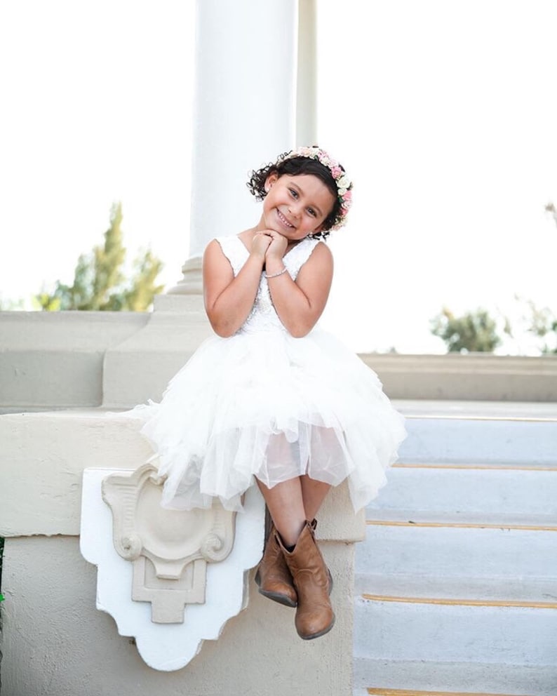 May include: A young girl wearing a white dress and brown boots sits on a white step with a flower crown in her hair. She is smiling and looking at the camera.