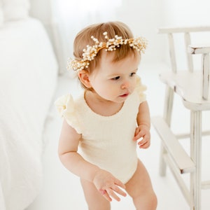 Puede incluir: Una ni&ntilde;a peque&ntilde;a con un body blanco con volantes y una corona de flores doradas con perlas blancas. La ni&ntilde;a est&aacute; de pie frente a una silla de madera blanca.