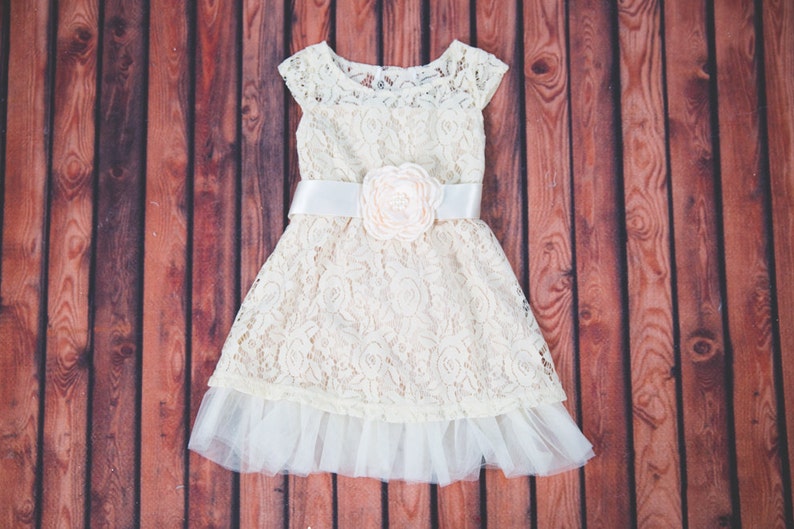 Peut inclure: Une robe en dentelle blanche avec un ruban en satin blanc et une grande fleur blanche en accent. La robe a une couche inf&eacute;rieure en tulle.