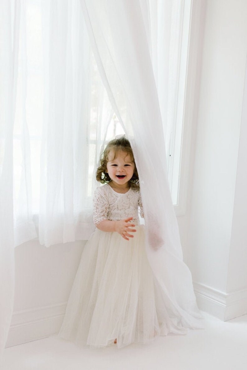 May include: A young child wearing a white lace bodice dress with a tulle skirt, smiling and peeking out from behind a sheer white curtain. The room is brightly lit with natural light.