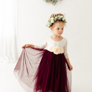May include: A young girl wearing a white lace dress with a burgundy tulle skirt and a flower crown. She is holding the skirt with one hand and looking down.