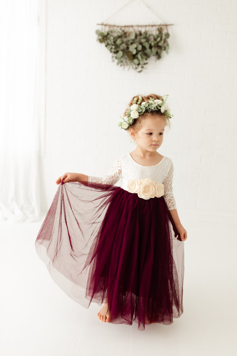 May include: A young girl wearing a white lace dress with a burgundy tulle skirt and a flower crown. She is holding the skirt with one hand and looking down.