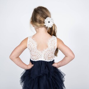 Puede incluir: Una ni&ntilde;a peque&ntilde;a con un corpi&ntilde;o de encaje blanco y una falda de tul azul marino de varias capas. El corpi&ntilde;o tiene la espalda baja y la falda est&aacute; fruncida en la cintura. La ni&ntilde;a tiene el pelo rubio con una pinza de flor blanca en el pelo.