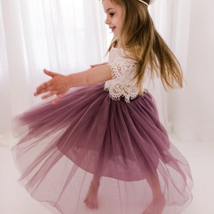 May include: A young girl wearing a white lace top and a mauve tulle skirt. She is twirling and smiling. The skirt is full and flowing, and the top has a delicate lace design.