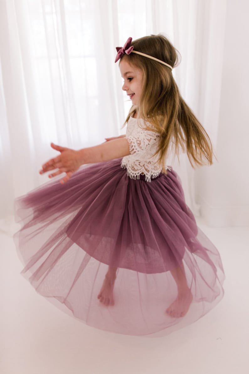 May include: A young girl wearing a white lace top and a mauve tulle skirt. She is twirling and smiling. The skirt is full and flowing, and the top has a delicate lace design.