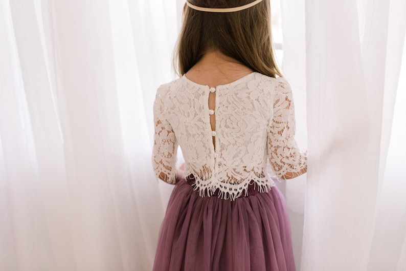 May include: A two-piece outfit consisting of a white lace top with three-quarter length sleeves, a scalloped hem, and a back button closure with pearl-like buttons, paired with a floor-length, gathered, solid mauve tulle skirt with a waistband.