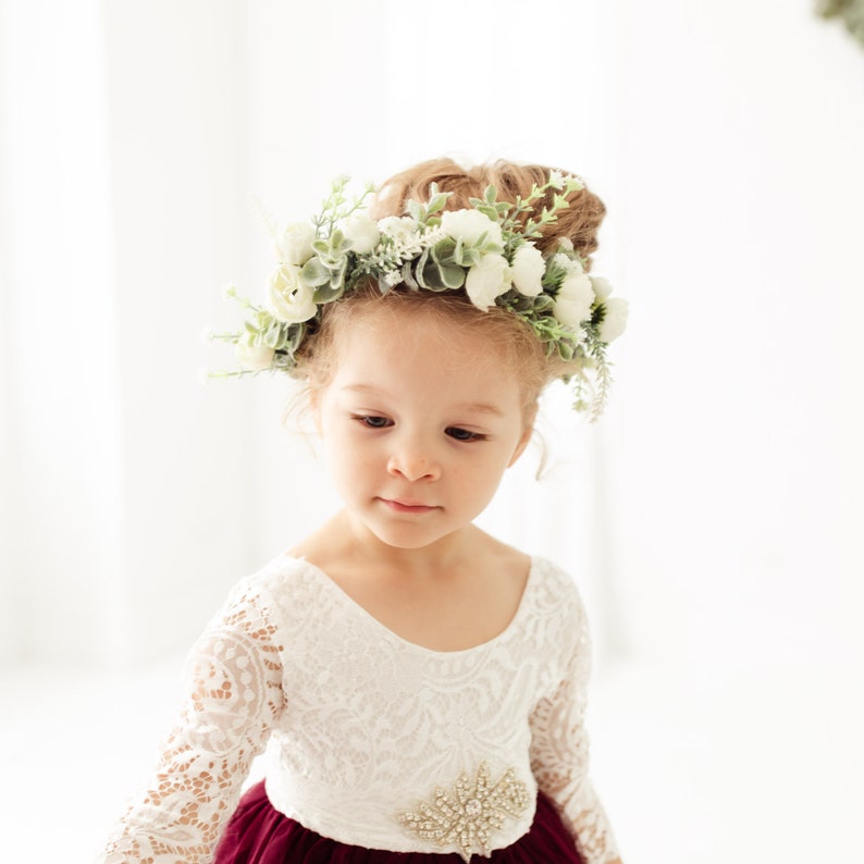 May include: A young girl wearing a white lace dress with a burgundy skirt and a flower crown. The crown is made of white flowers and green leaves.