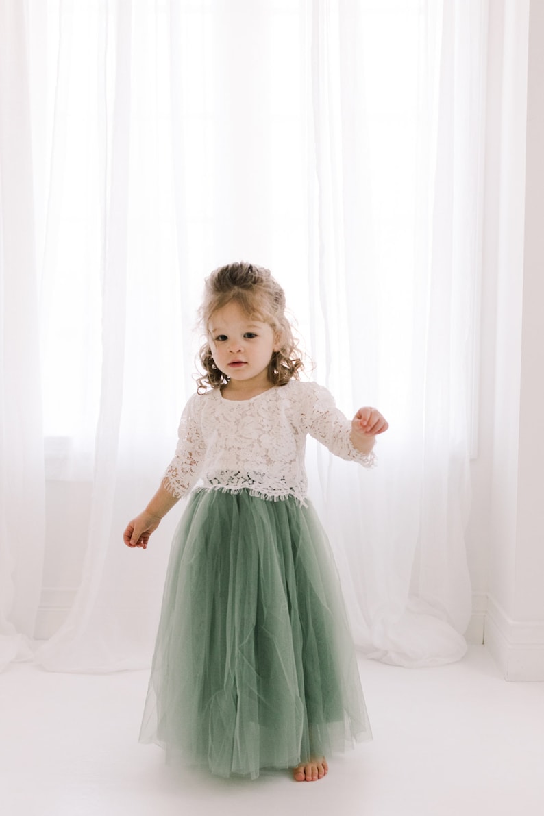 May include: A young girl wearing a white lace top and a sage green tulle skirt. She is standing in front of a white curtain.