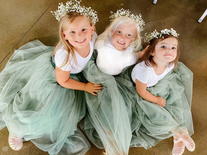 May include: Three young girls wearing white shirts and sage green tulle skirts with flower crowns. The girls are sitting on a light brown floor.