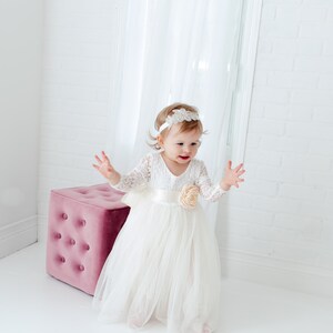 May include: A white long-sleeved dress featuring a lace bodice, a satin waistband with a large fabric flower accent, and a layered tulle skirt. The outfit is paired with a white headband adorned with a decorative silver-toned crystal embellishment.