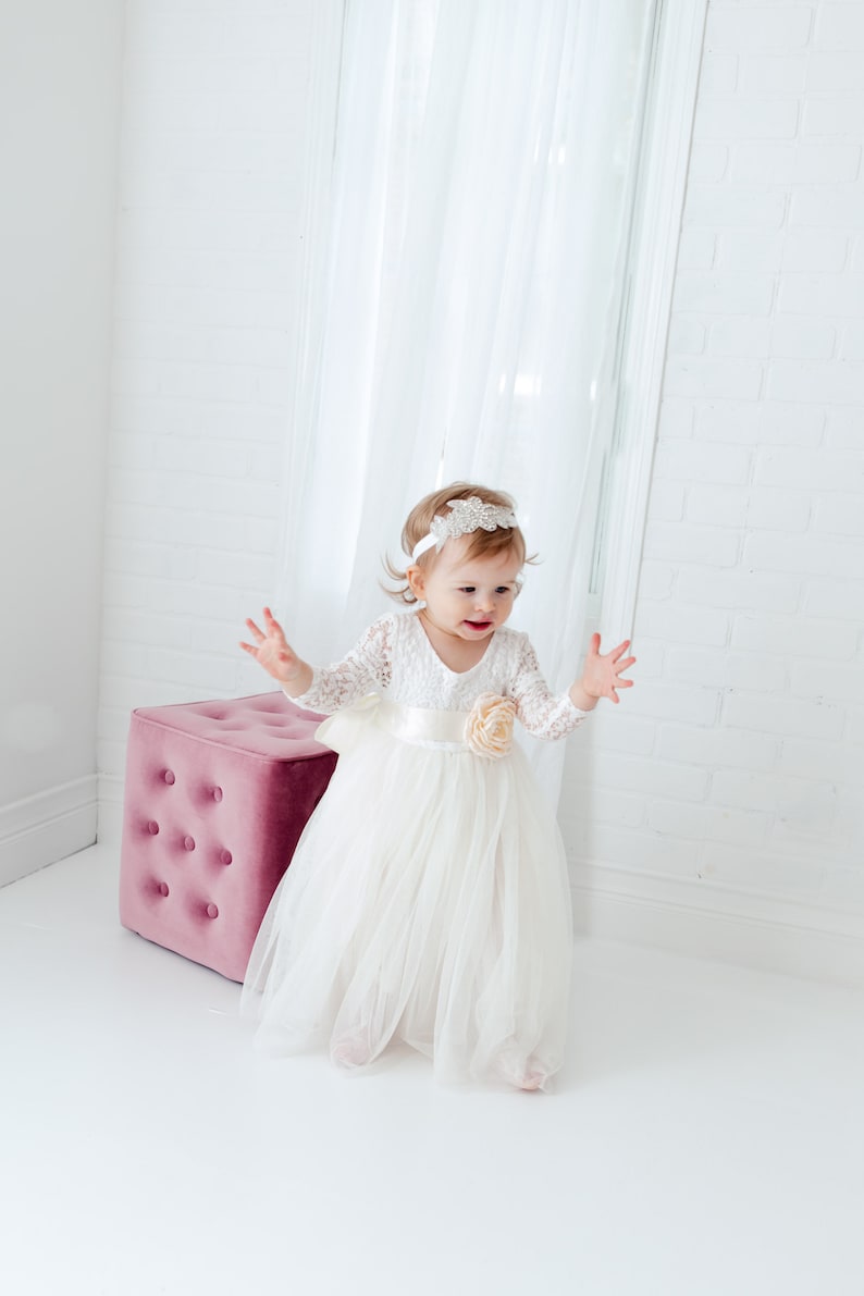 May include: A white long-sleeved dress featuring a lace bodice, a satin waistband with a large fabric flower accent, and a layered tulle skirt. The outfit is paired with a white headband adorned with a decorative silver-toned crystal embellishment.