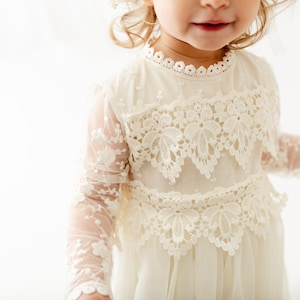 May include: An off-white dress featuring long sheer sleeves with floral embroidery, a high neckline with scalloped lace trim, and two horizontal bands of intricate floral lace appliqu&eacute; across the bodice, set against a plain white background.