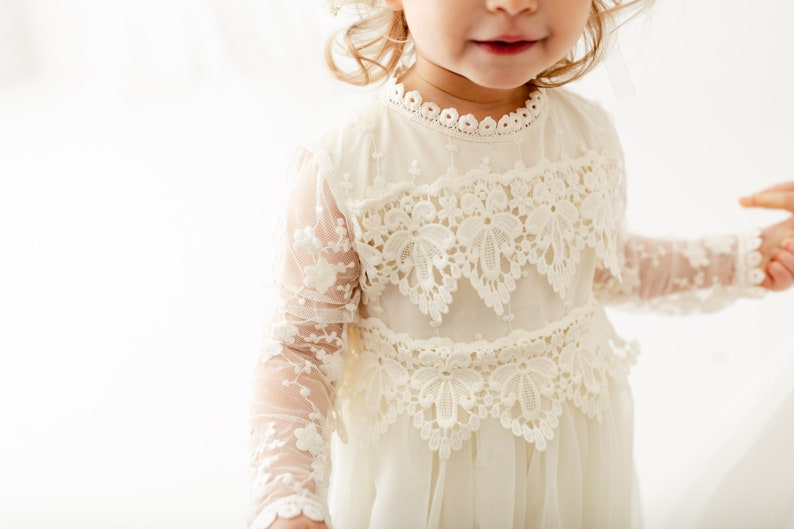 May include: An off-white dress featuring long sheer sleeves with floral embroidery, a high neckline with scalloped lace trim, and two horizontal bands of intricate floral lace appliqu&eacute; across the bodice, set against a plain white background.