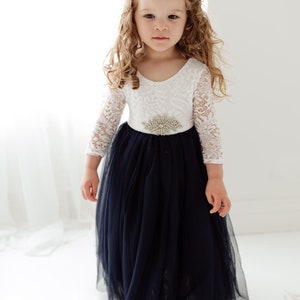May include: A young girl wearing a white lace long-sleeved dress with a navy blue tulle skirt and a jeweled belt.