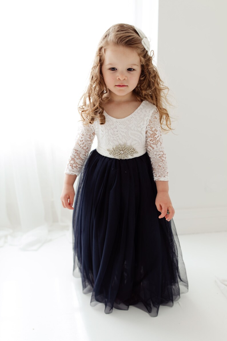 May include: A young girl wearing a white lace long-sleeved dress with a navy blue tulle skirt and a jeweled belt.