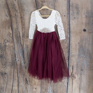 May include: A white lace dress with long sleeves and a burgundy tulle skirt. The dress has a jeweled belt around the waist.